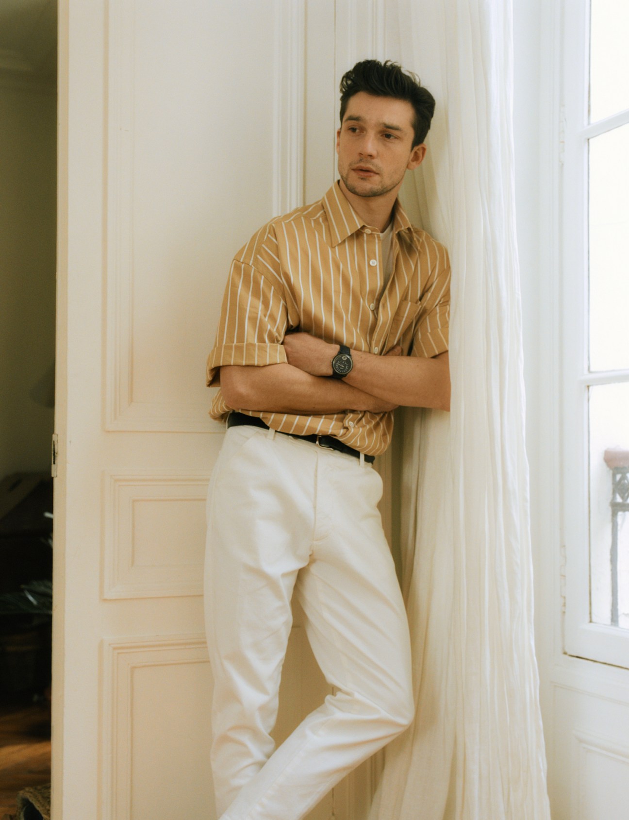 Man in tan striped shirt and cream trousers leaning against white panelled door.