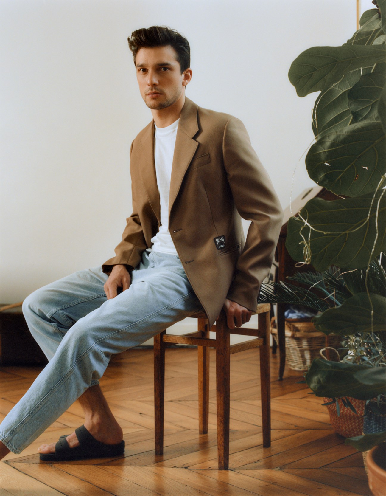 Man sitting on wooden chair wearing camel blazer, white t-shirt, light blue jeans and black sandals next to potted plant.