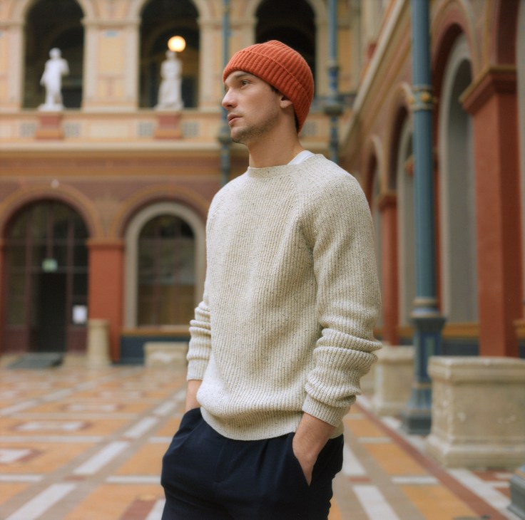 Man wearing cream APC jumper, navy trousers, and coral Begg & Co beanie in a courtyard with classical architecture.