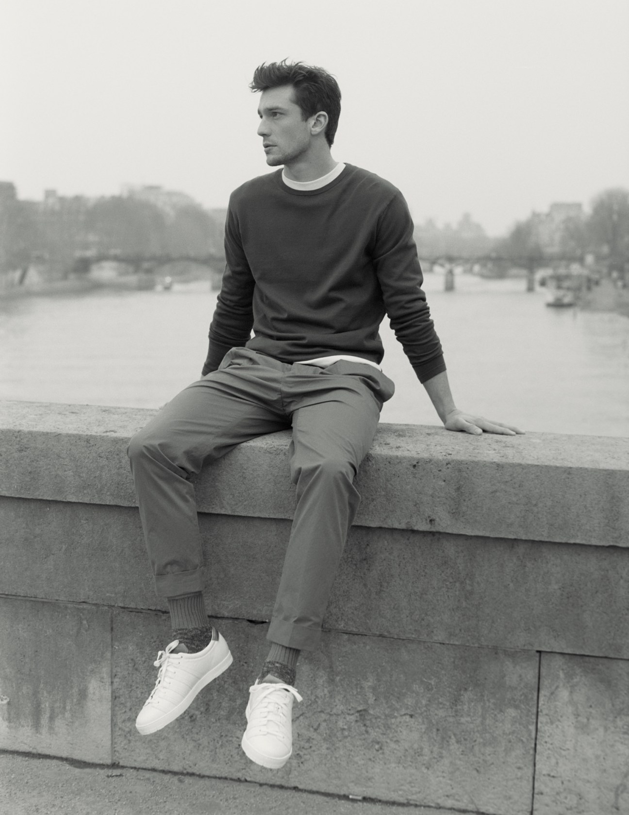 Man sitting on concrete wall overlooking water, wearing jumper and long-sleeve T-shirt by Sunspel, trousers by Officine Générale, socks by Golden Goose Deluxe Brand, trainers by Corthay.