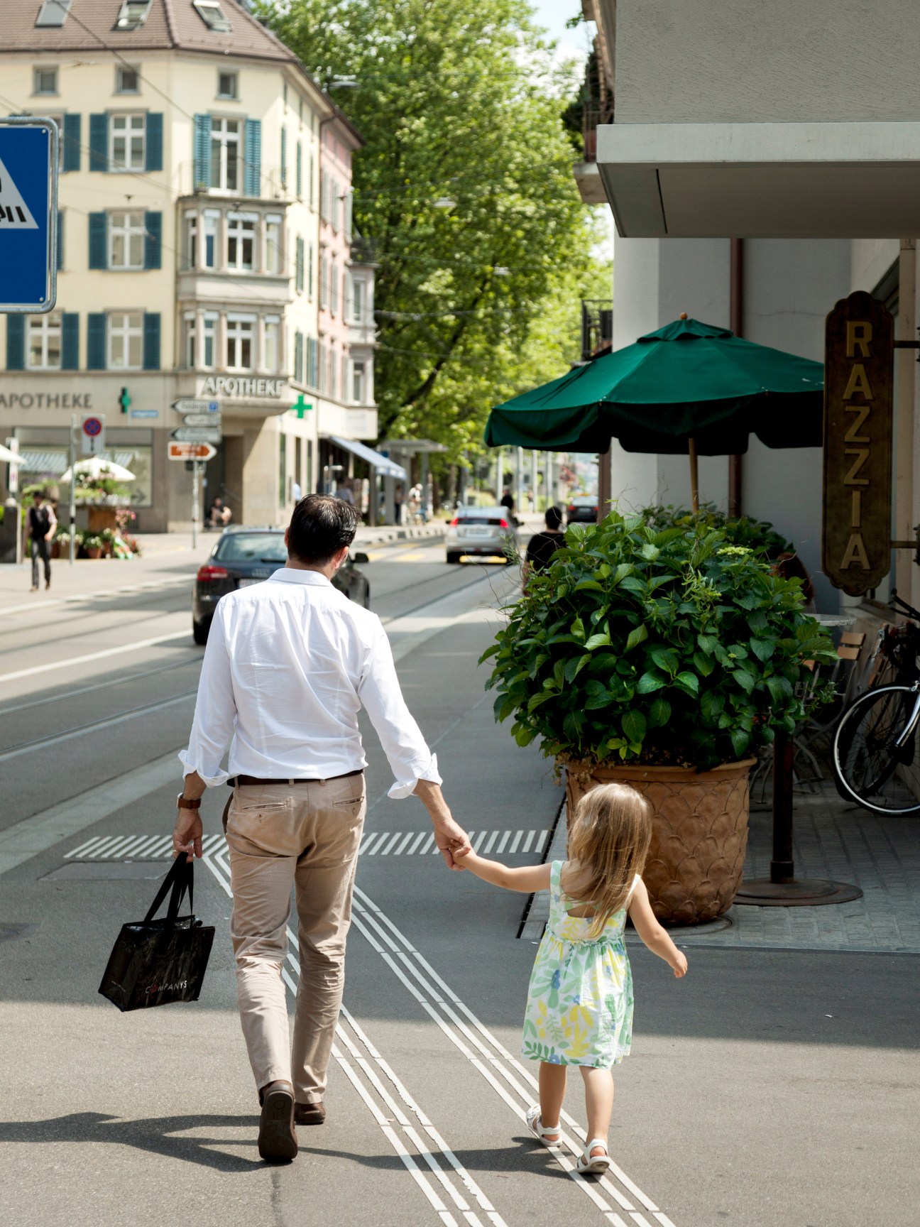 A man and young child walking hand-in-hand on a tree-lined Zurich street with outdoor café seating.