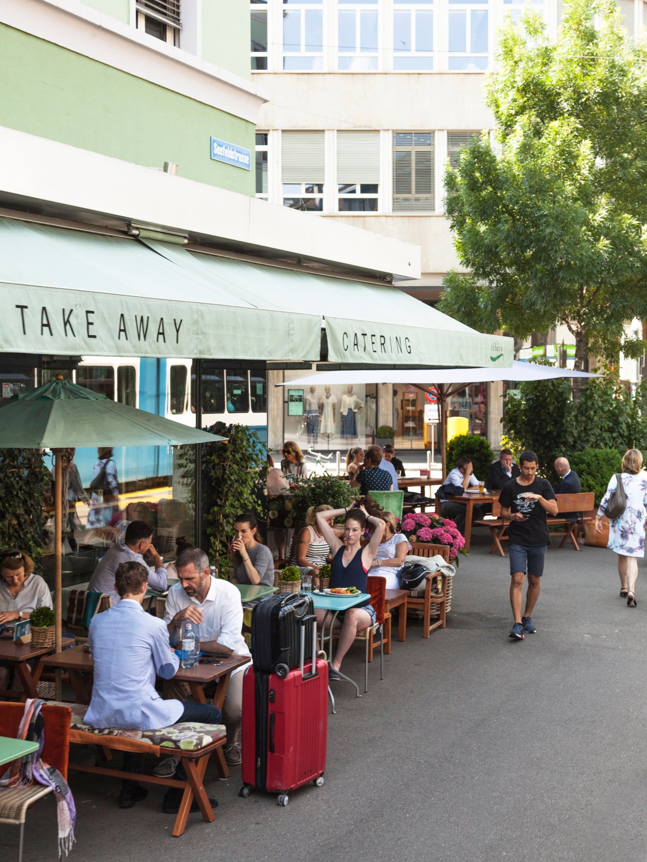 People dining outdoors at a street-side restaurant with green umbrellas and ivy-covered buildings in Zurich's Seefeld district.