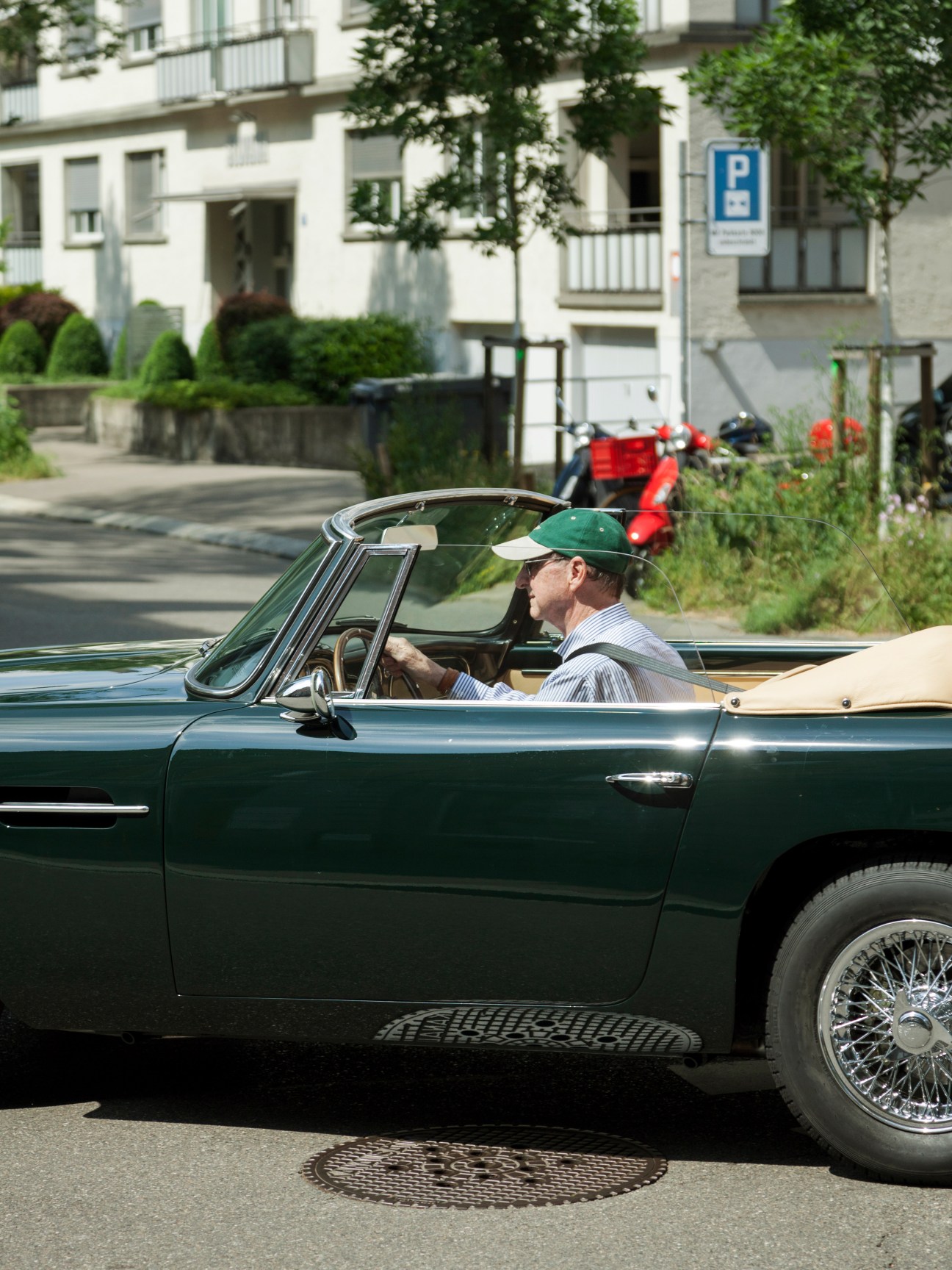 Man driving a vintage green convertible car on a Zürich street with residential buildings in background.
