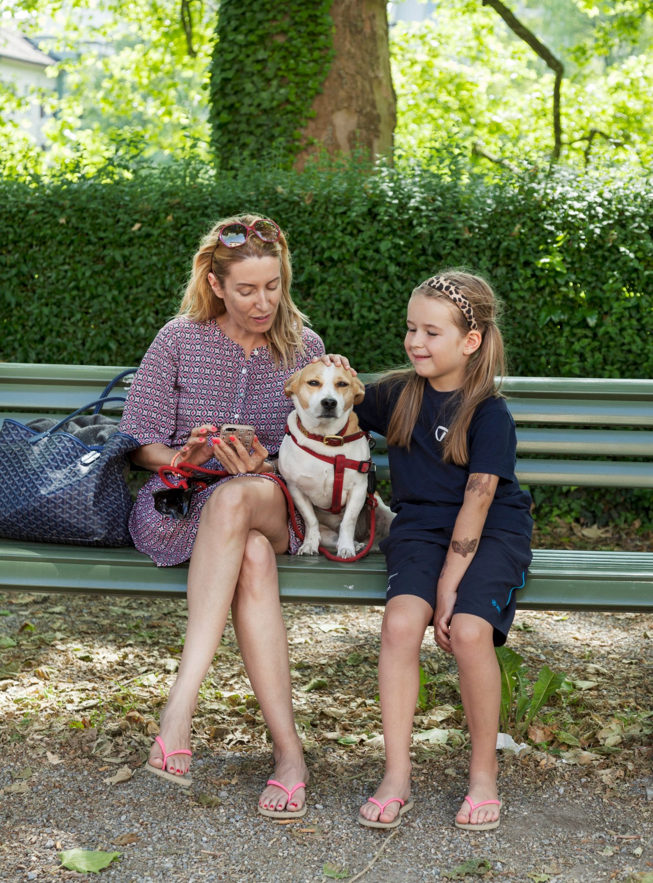 Woman and girl sitting on a bench with a dog in a park surrounded by ivy-covered trees.