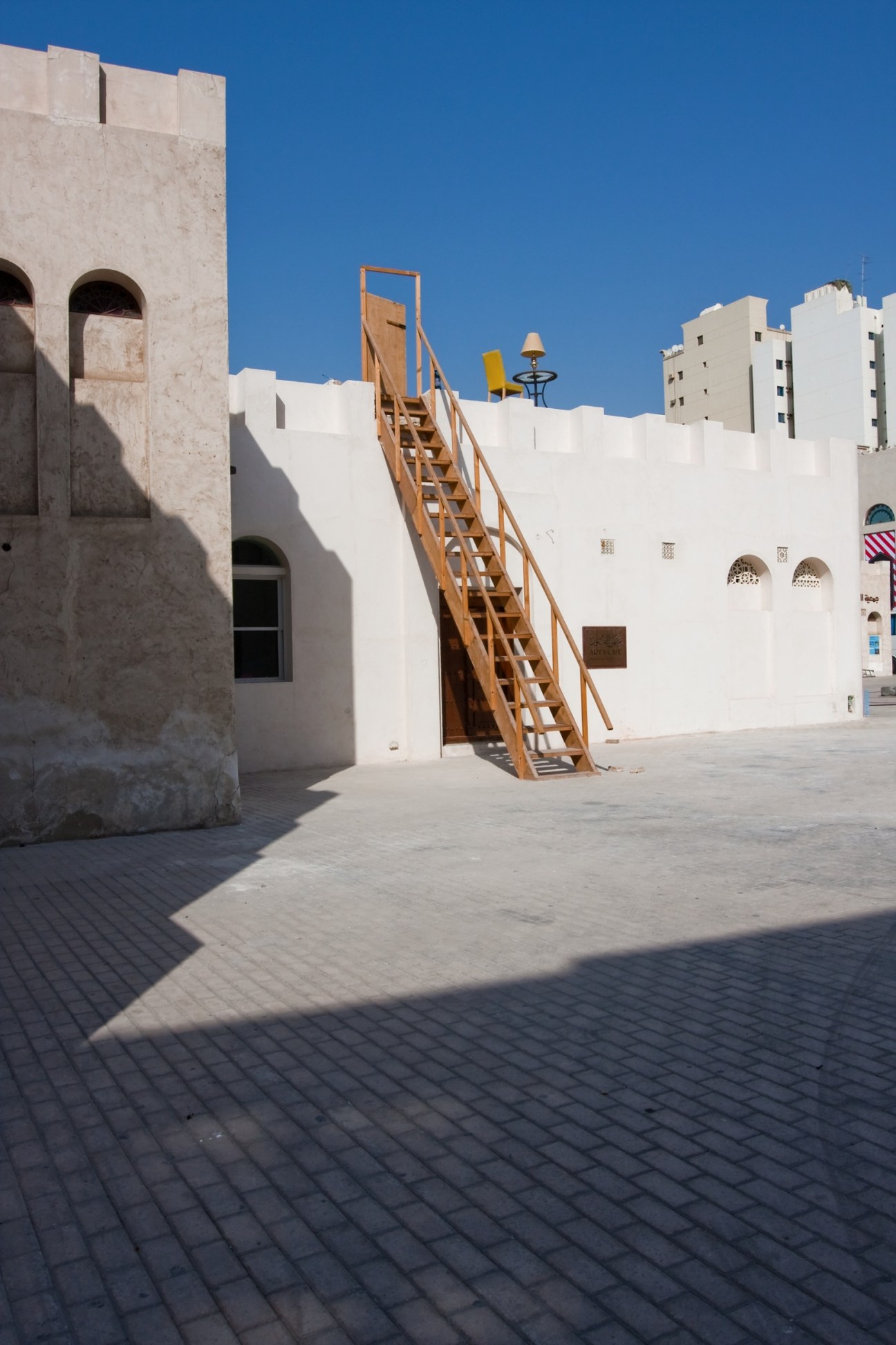 White-walled courtyard with wooden ladder installation against clear blue sky in Sharjah.