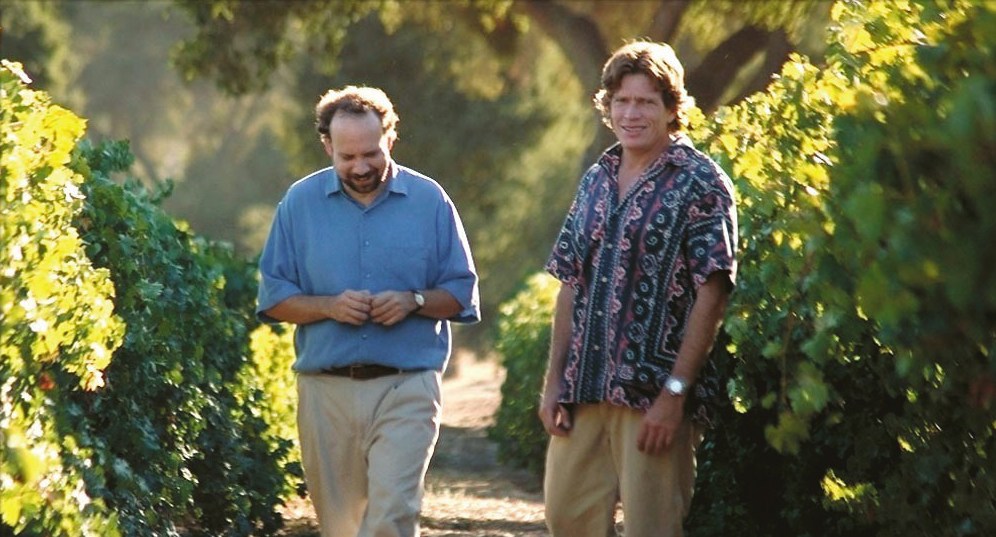 Two men walking through a vineyard lined with green grapevines in sunlight.