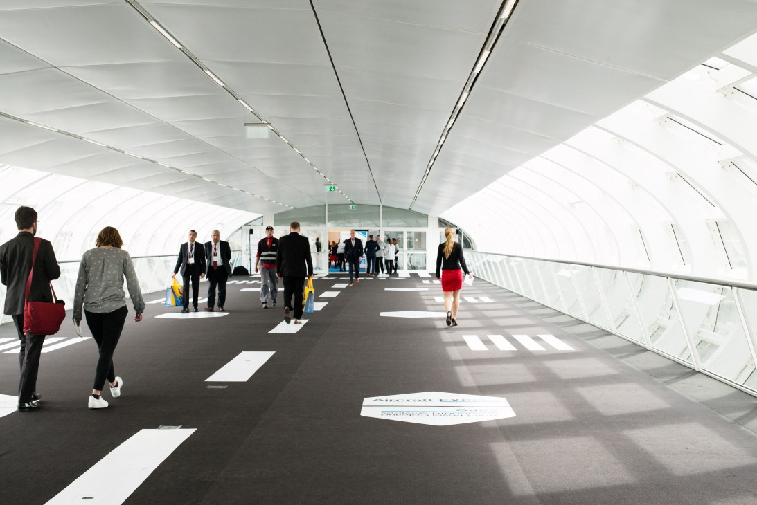 People walking through a modern white tunnel with runway-style markings on the floor.