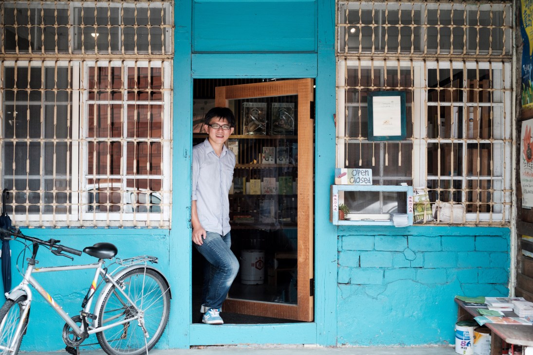 Stay Traveler Bookstore founder Jay Lin standing in doorway of turquoise storefront with bicycle parked outside