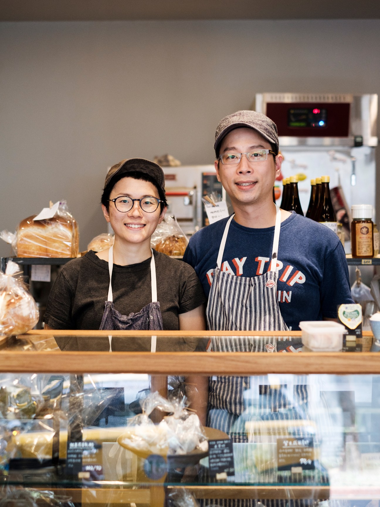 Two smiling staff members wearing aprons and glasses standing behind a bakery display case at Casan bakery