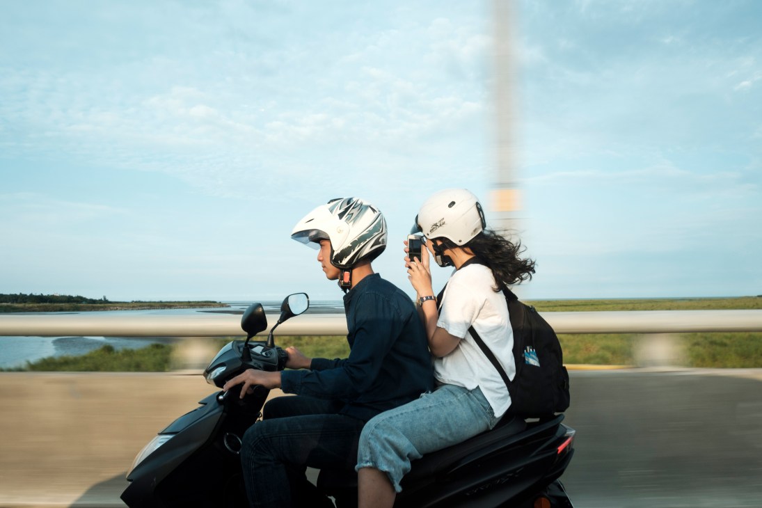 Two people wearing helmets riding a motorcycle on a bridge with scenic landscape in background