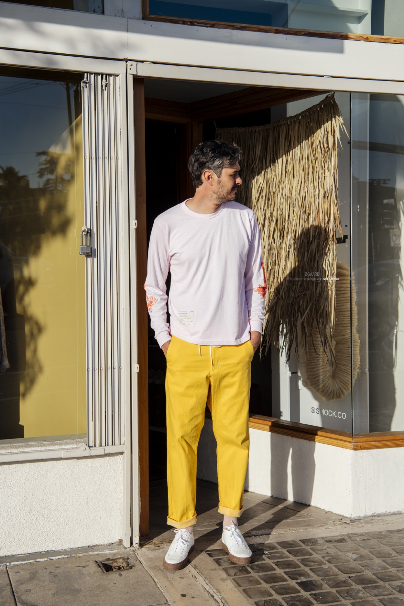 Man wearing Smock brand lavender long-sleeve shirt and yellow cropped trousers outside a storefront.