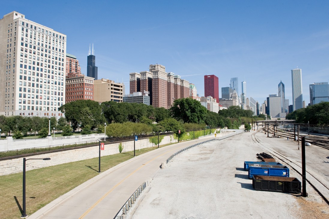 Chicago skyline viewed from a lakeside path with green spaces, buildings, and moored boats in foreground.
