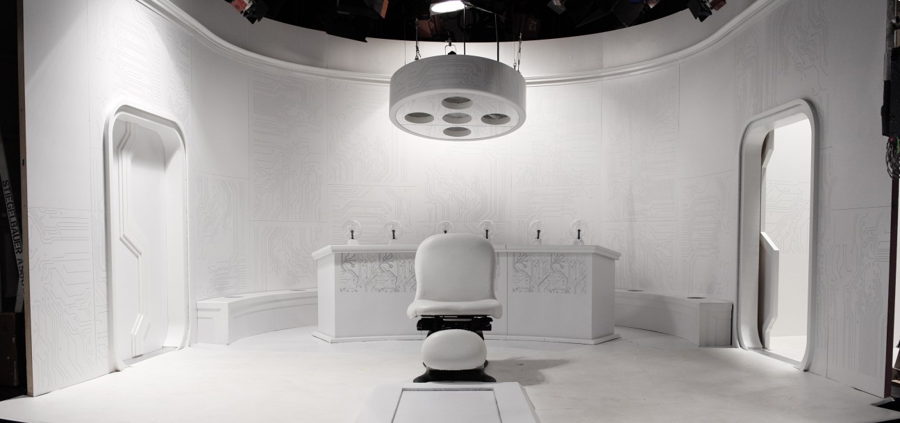 A futuristic white spaceship interior set with curved walls, overhead lighting, and a central control station for Saturday Night Live.