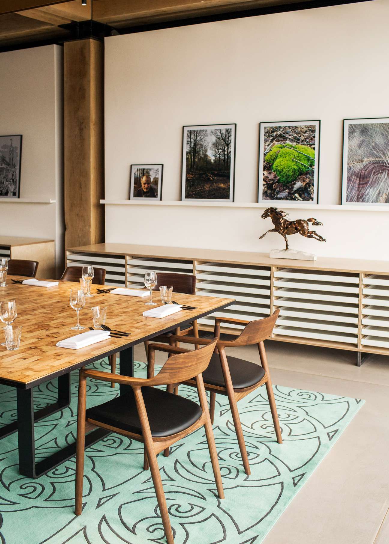 Rooftop meeting room with wooden table, walnut chairs, patterned turquoise rug, and framed photographs on white wall shelving.