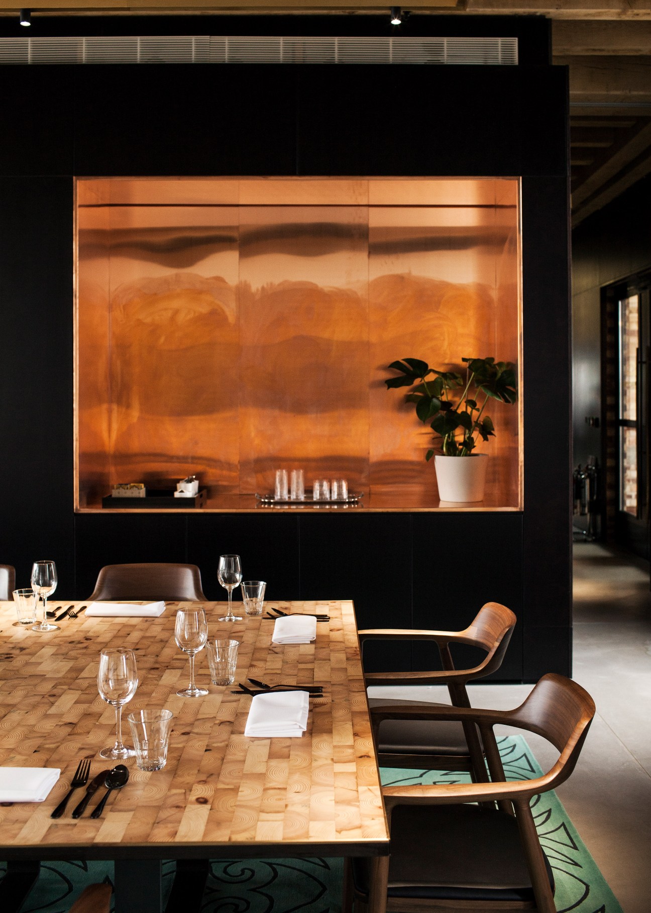 Modern dining table with wooden chairs and place settings below an illuminated copper-clad display box on black wall.