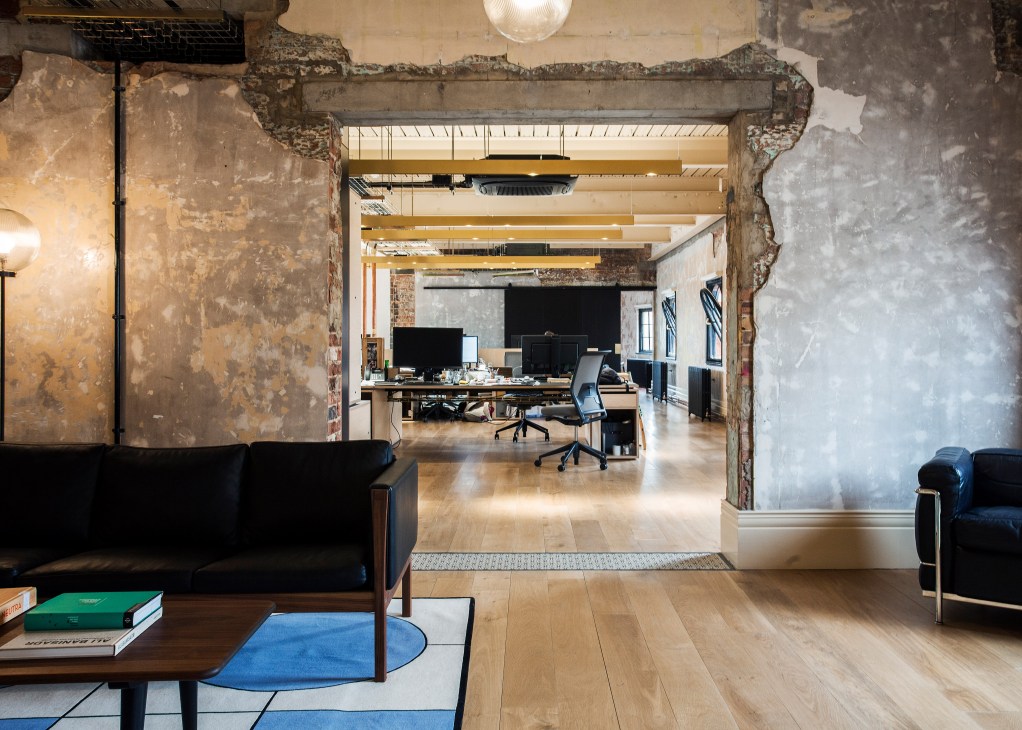 Open-plan office interior with exposed brick walls, wooden beams, and modern workstations visible through deteriorated doorway.