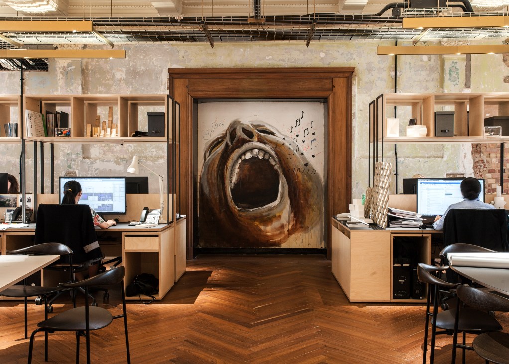 Open office with wooden workstations flanking a large wooden-framed mural of a screaming face with musical notes on an exposed brick wall.