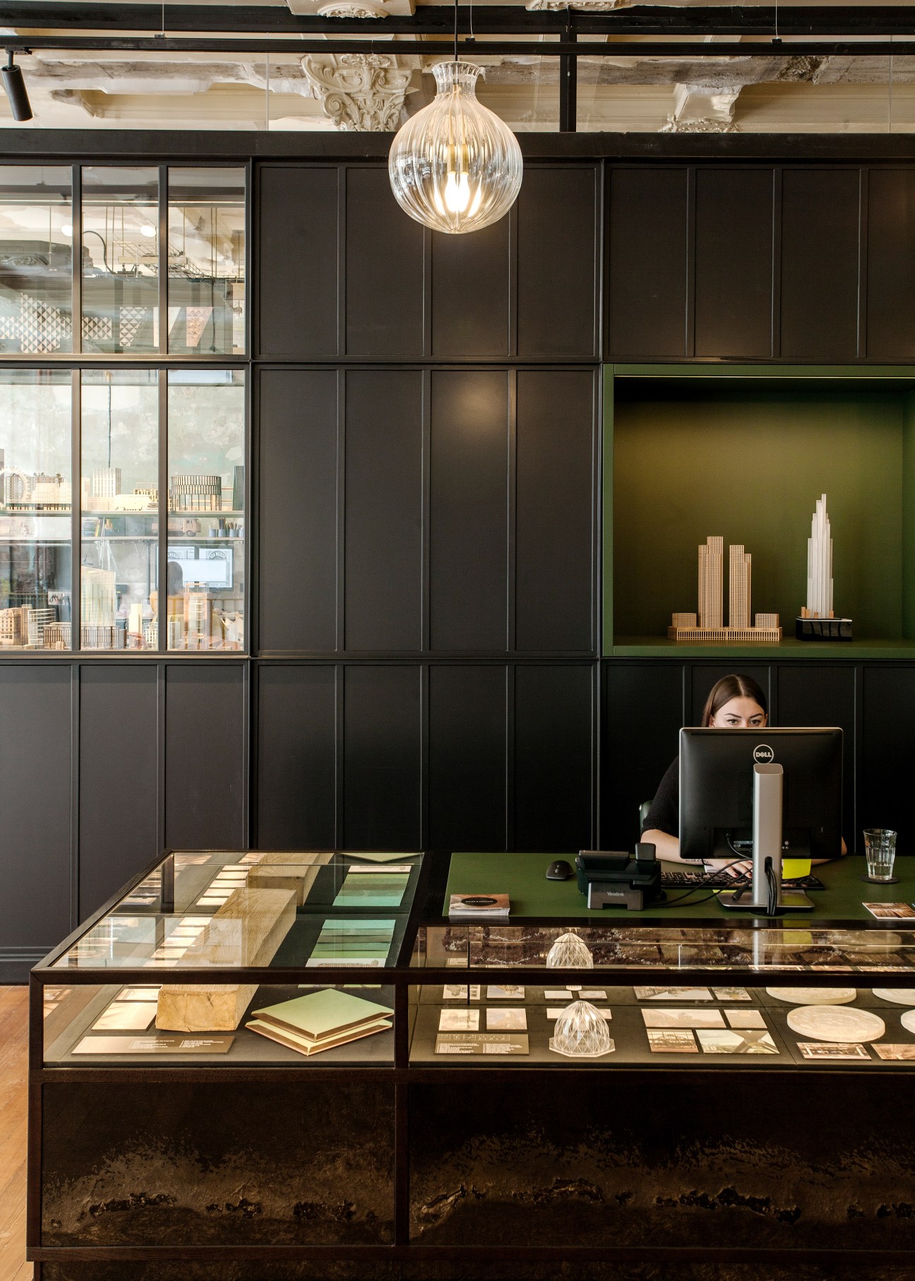 Modern office reception desk with black panelling, glass display cases, hanging Lasvit glass pendant light, and staff member working at computer.