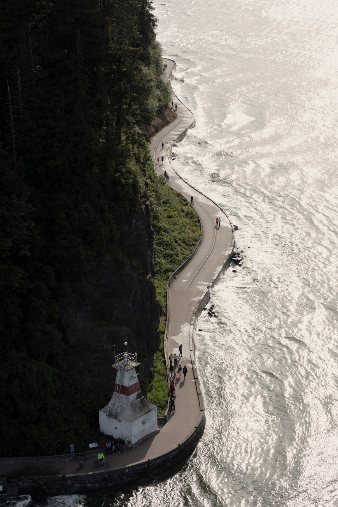 Aerial view of curved waterfront pathway with lighthouse, people walking along shore next to forested coastline