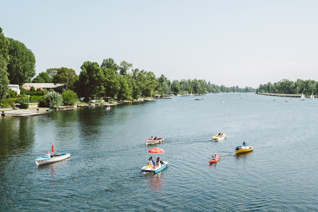 People in colorful boats on the Danube River with tree-lined riverbanks and sailboats in the distance.