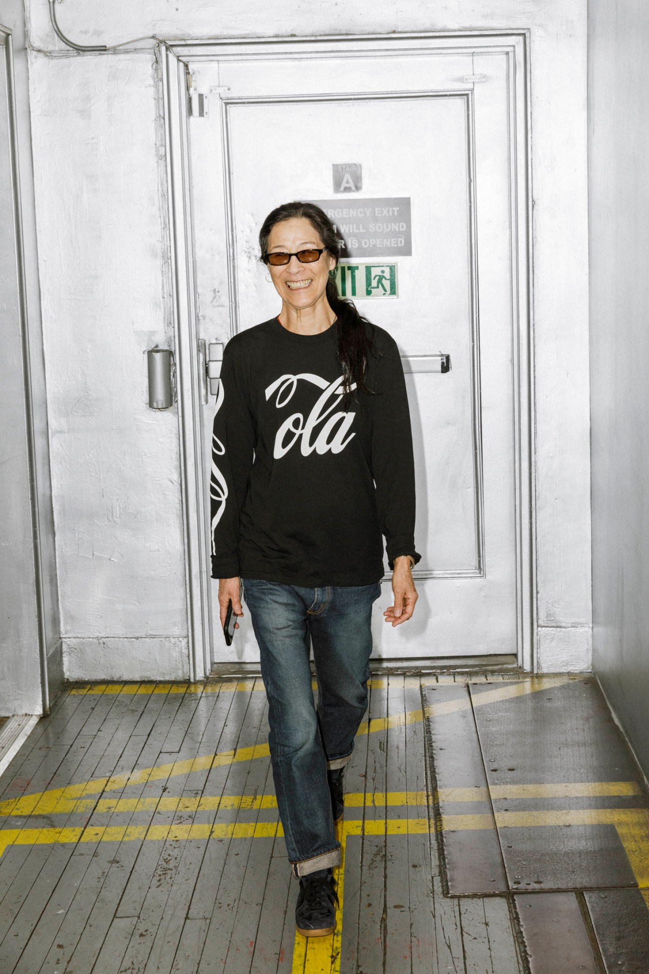 Stella Ishii standing in front of an emergency exit door wearing sunglasses, black sweater and jeans