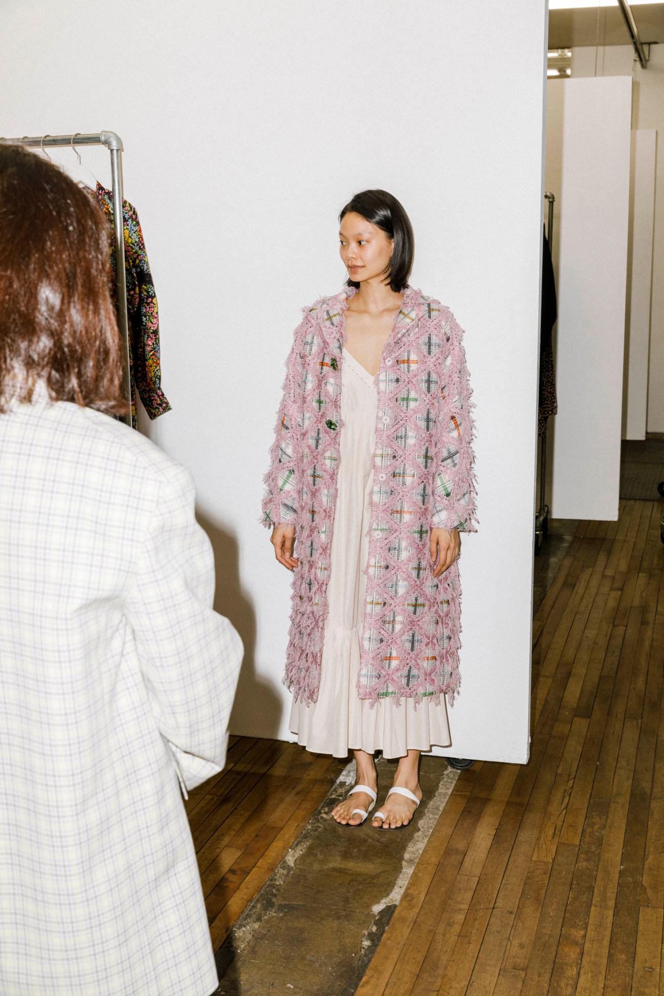 Woman wearing pink textured coat over white dress standing in showroom with clothing racks