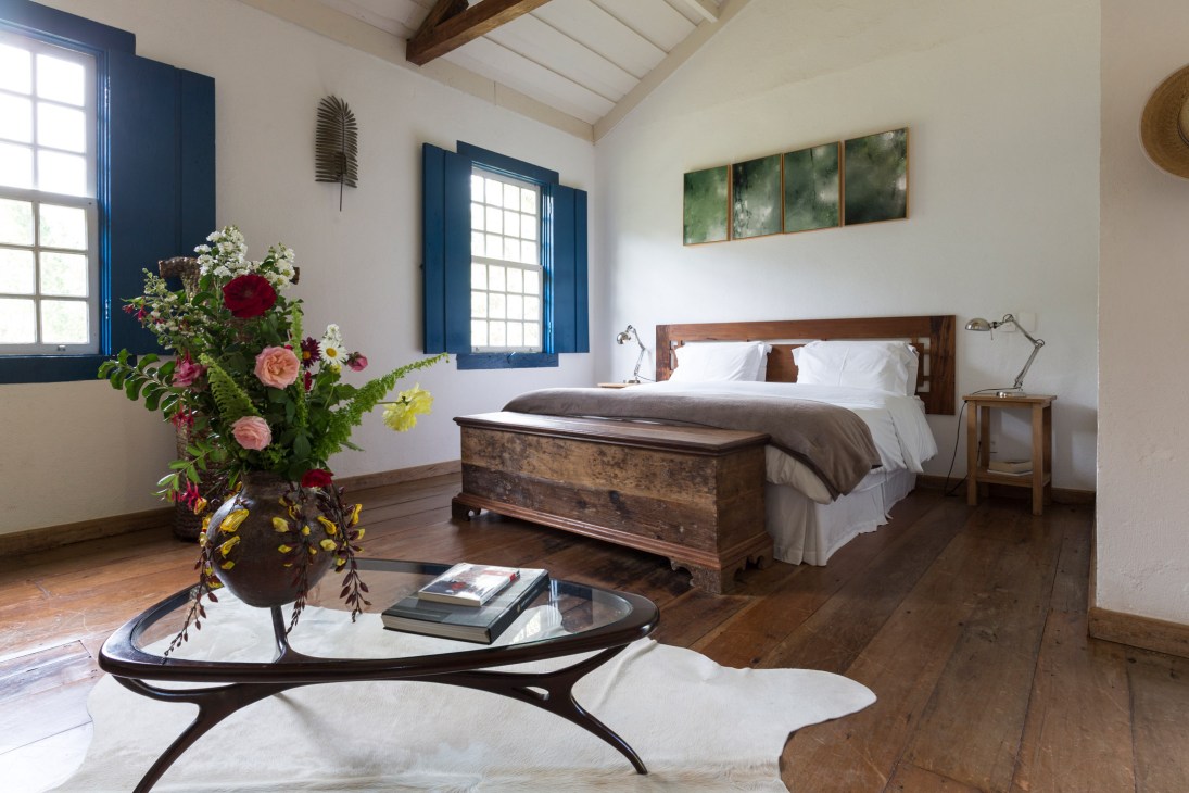 Bright bedroom with wooden bed, white bedding, blue-framed windows, and colorful flowers in vase on side table.