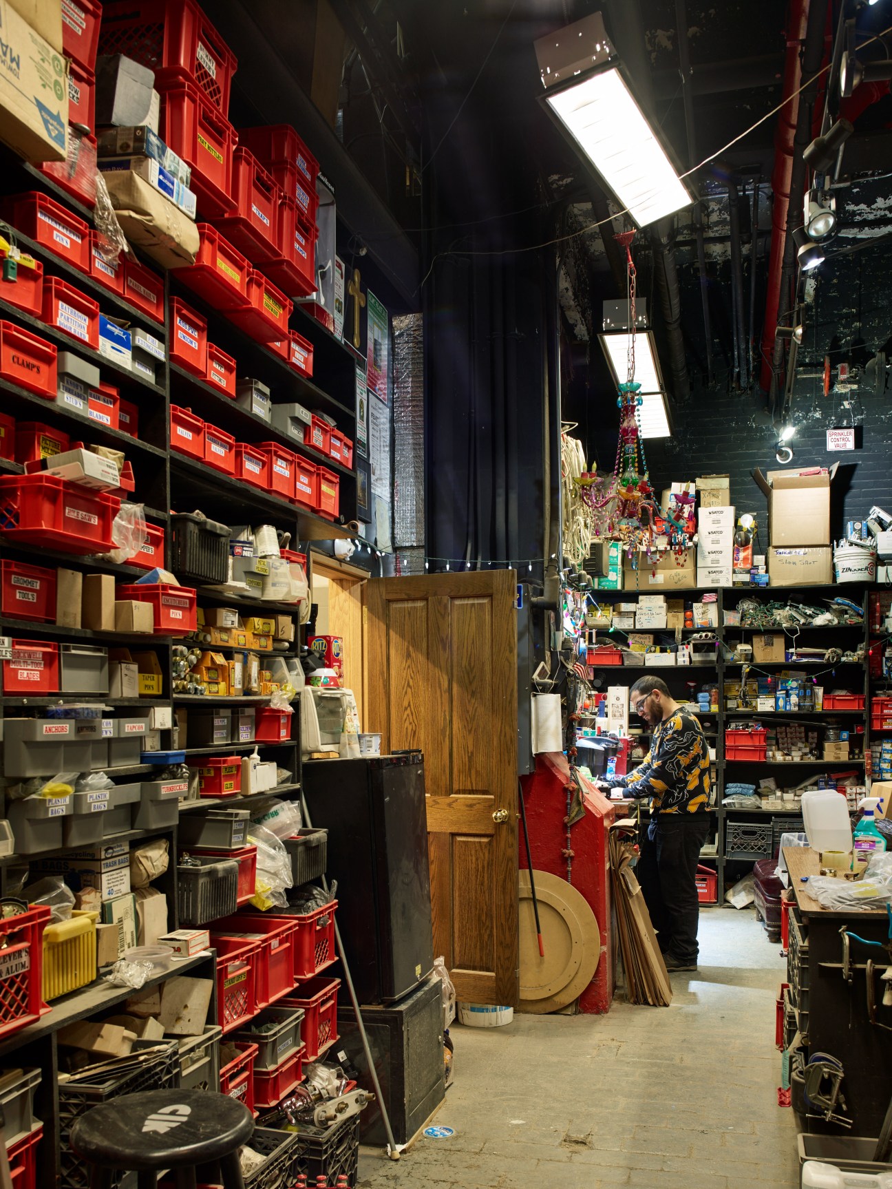 A worker in a patterned jacket stands in a cluttered supply room at Stiegelbauer Associates with red storage bins and shelves stacked with materials and props.
