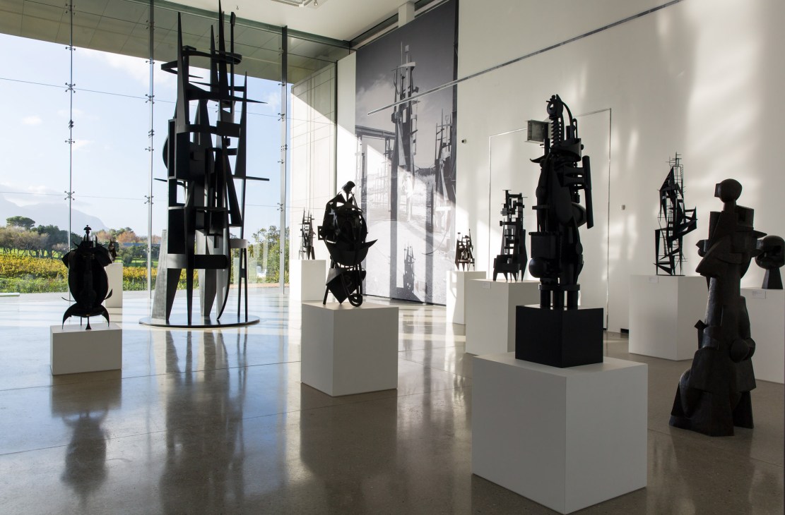 Modern art gallery exhibition featuring black sculptural installations on white pedestals with large windows overlooking vineyards.