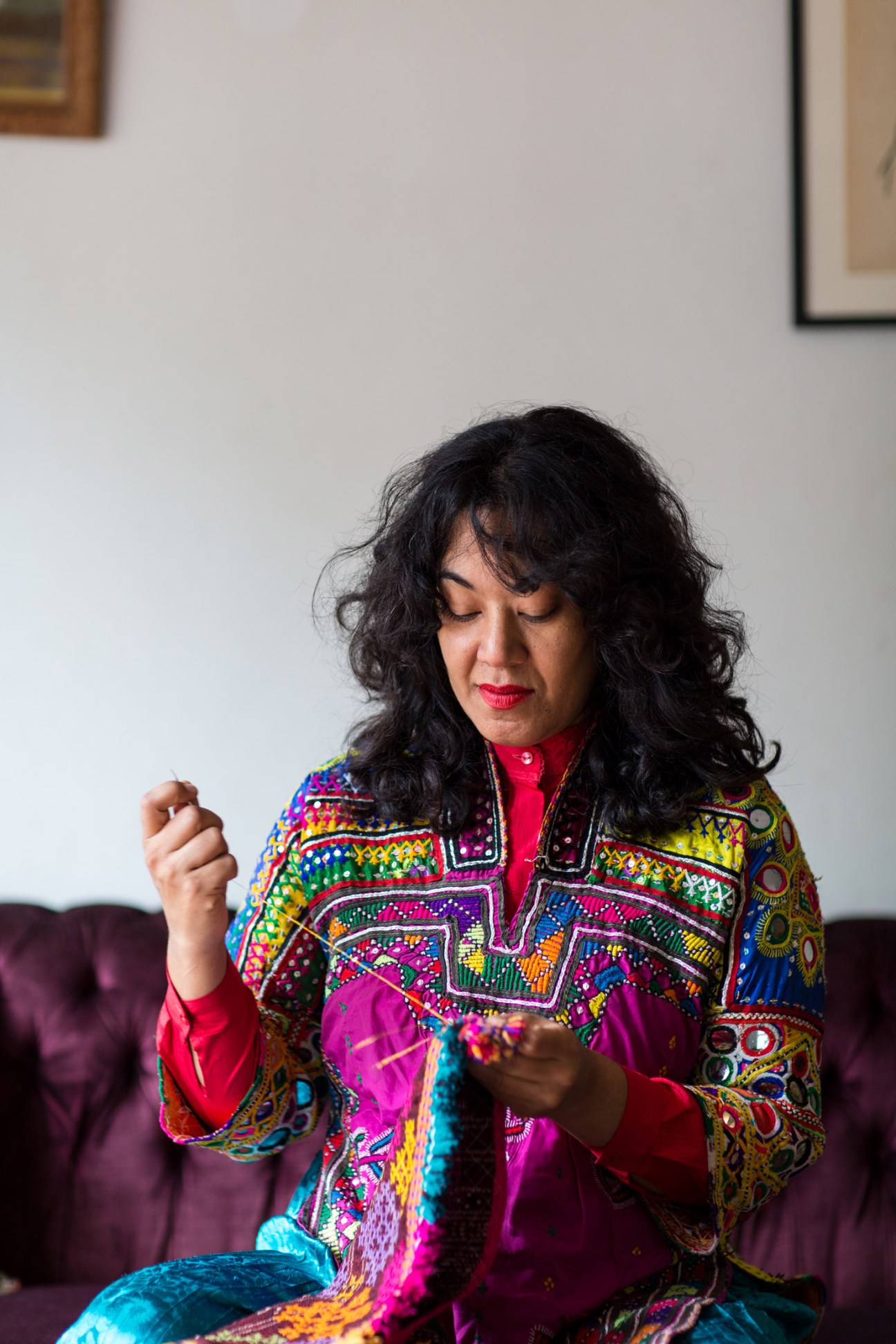 Thania Peterson, a visual artist, sits wearing a vibrant multicolored embroidered jacket over a red shirt, looking down at colorful textile work in her hands.