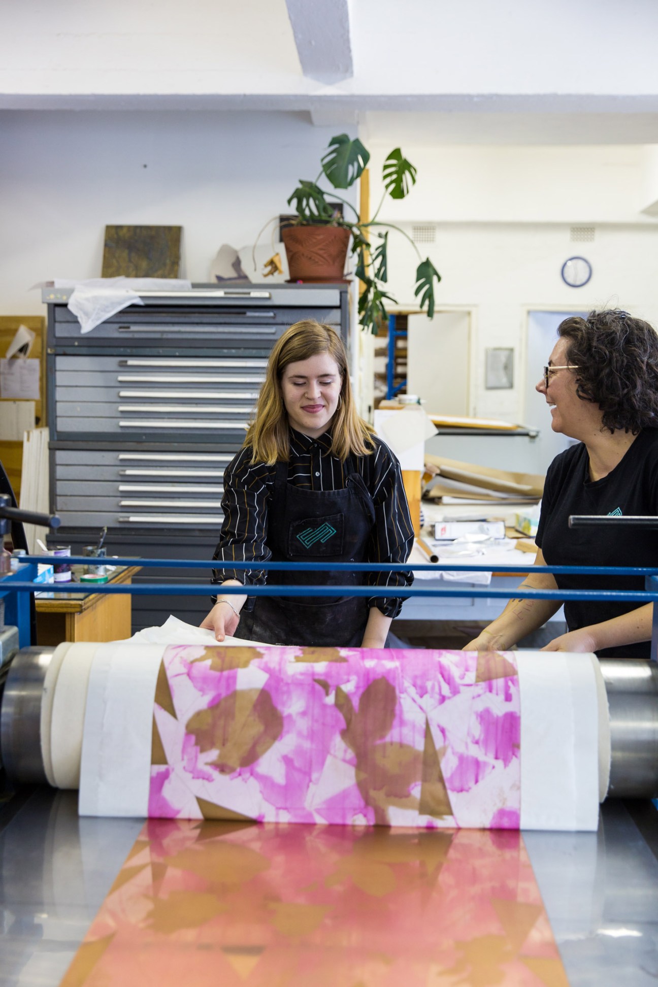 Two people examine a colorful printed fabric in a printmaking studio workspace.