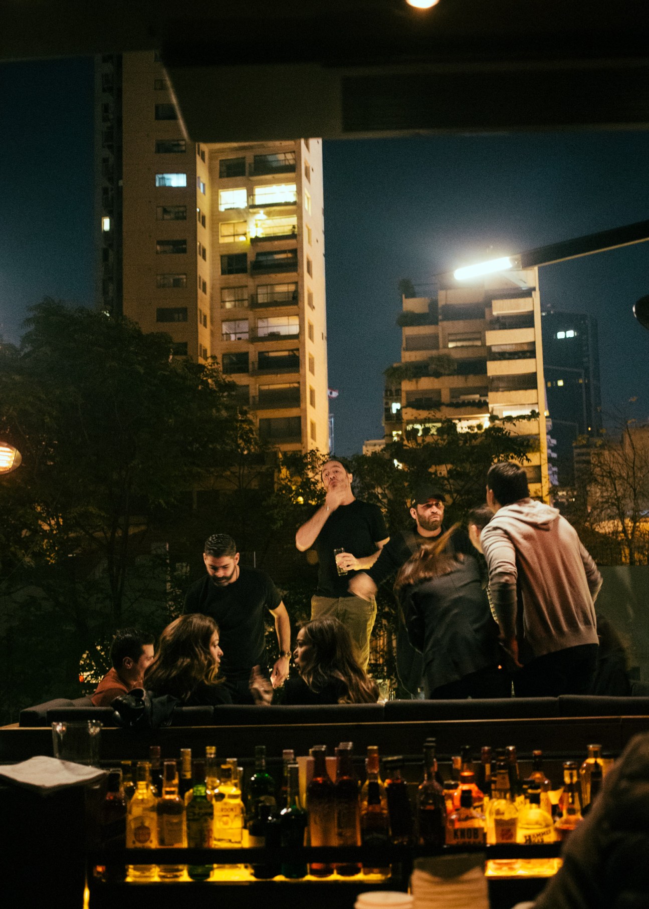 People socializing at an outdoor bar area at night with illuminated bottles displayed on a counter.