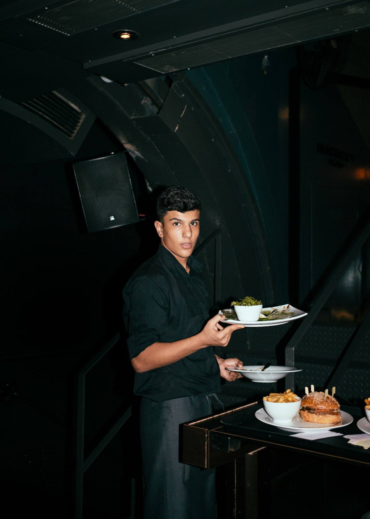 A waiter in black shirt carries empty plates in a dimly lit bar interior.