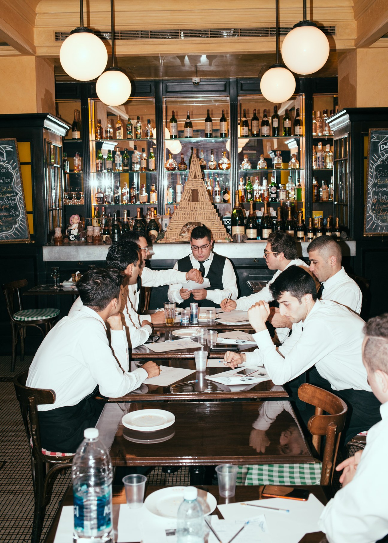 Waiters in white shirts and vests stand at a table in a modern bar with illuminated globe lights and a full bottle display behind them.
