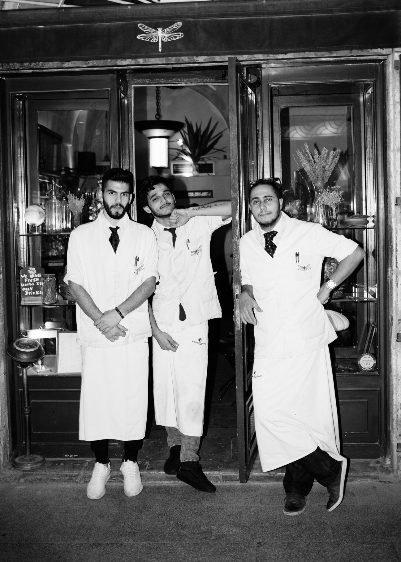 Three men in white lab coats and ties stand outside Dragonfly bar with a dragonfly logo above the entrance.