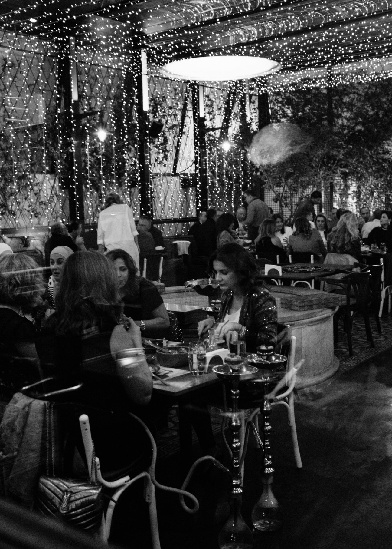 Diners gathered around tables at an upscale Lebanese restaurant with shisha pipes on the table and decorative string lights overhead.