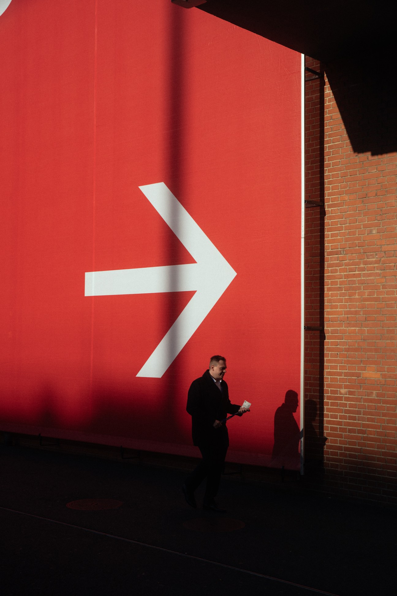 A man in dark clothing walking past a large red wall with a white directional arrow, next to a brick building