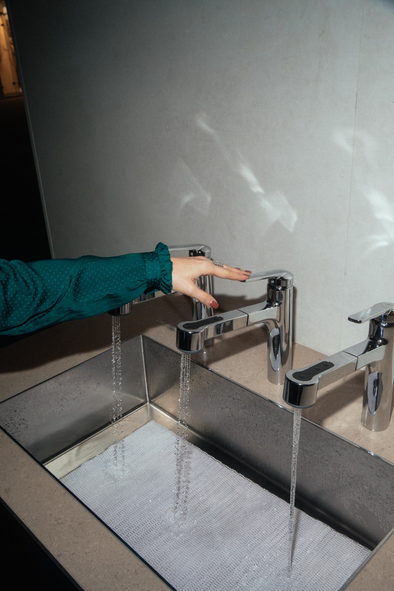 Person in teal sleeve activating touchless KWC sink fittings with water flowing into modern stainless steel basin