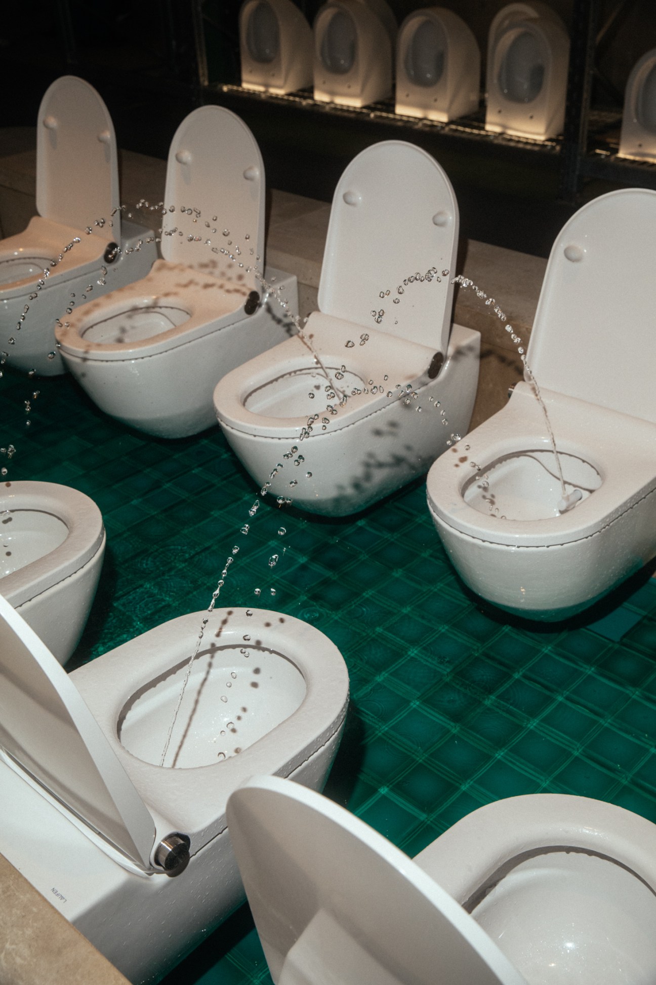 Multiple white Laufen toilets with water spraying from their bowls displayed on green tiled floor at trade show