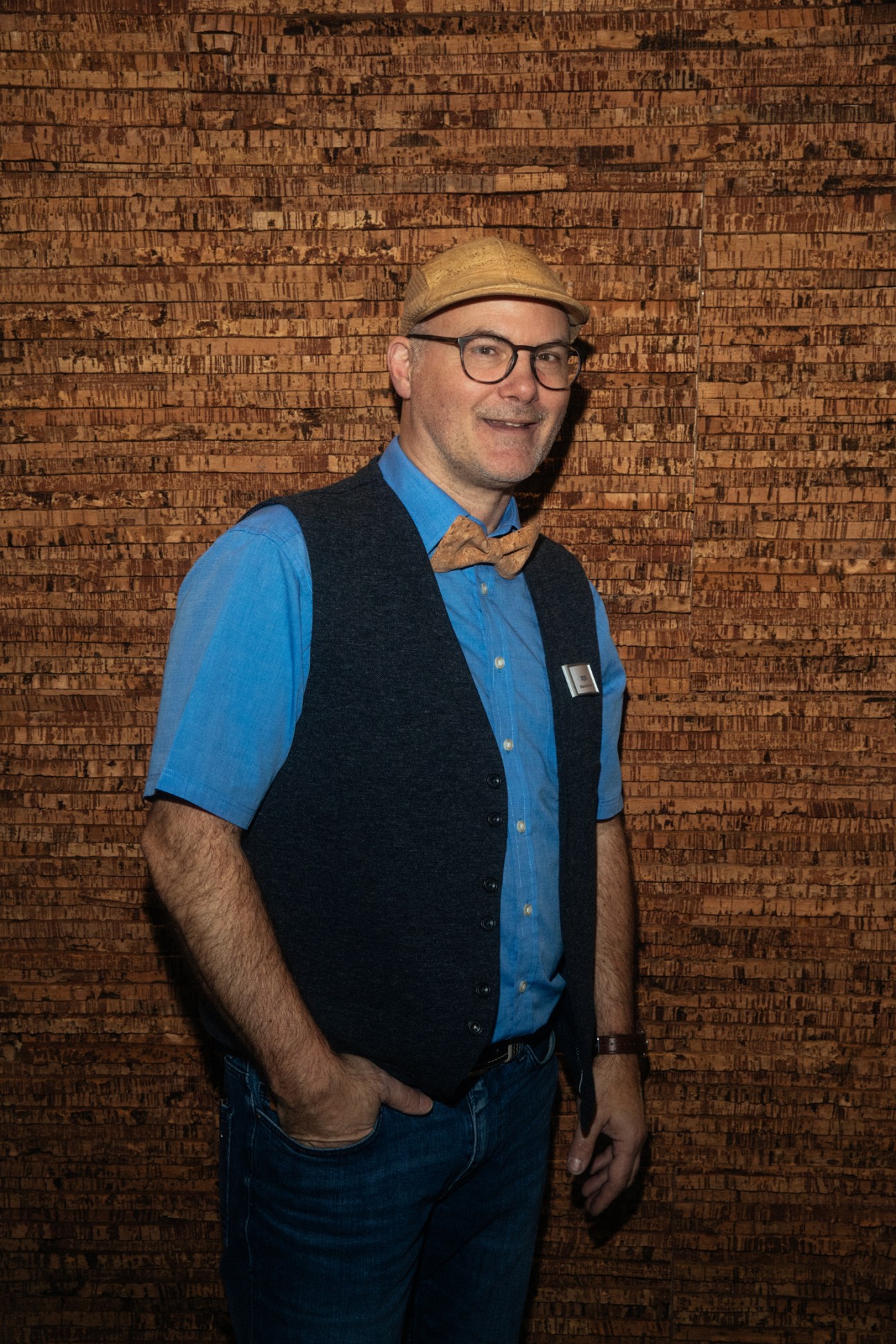 Man in hat, glasses, vest and bow tie standing in front of textured cork wall at trade fair