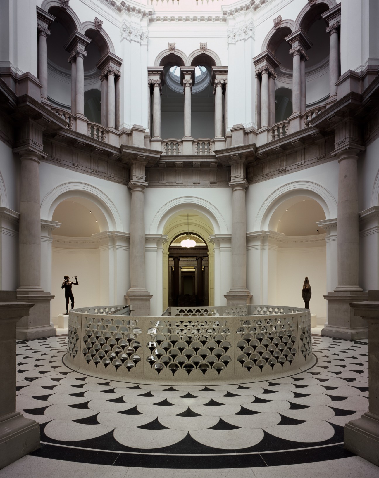 Spiral staircase with perforated metal railings in a neoclassical gallery rotunda with columns and skylights.