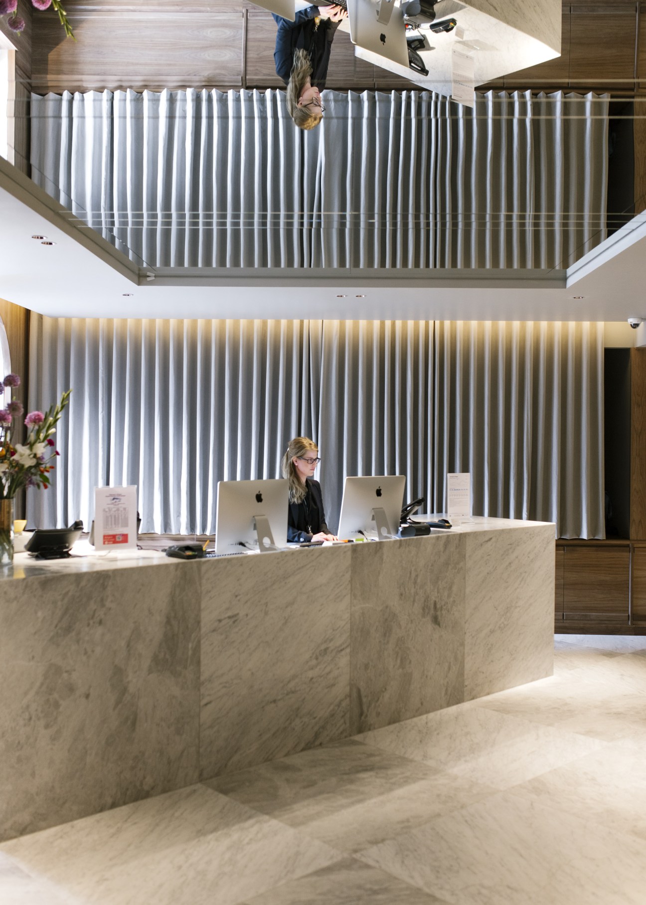 Modern hotel lobby with marble desk, vertical blinds, and staff member working at computers.
