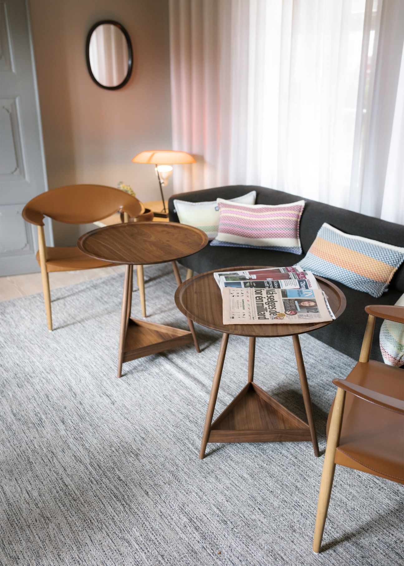 Modern lounge with wood furniture, colorful pillows on a dark sofa, and a round side table holding a newspaper.