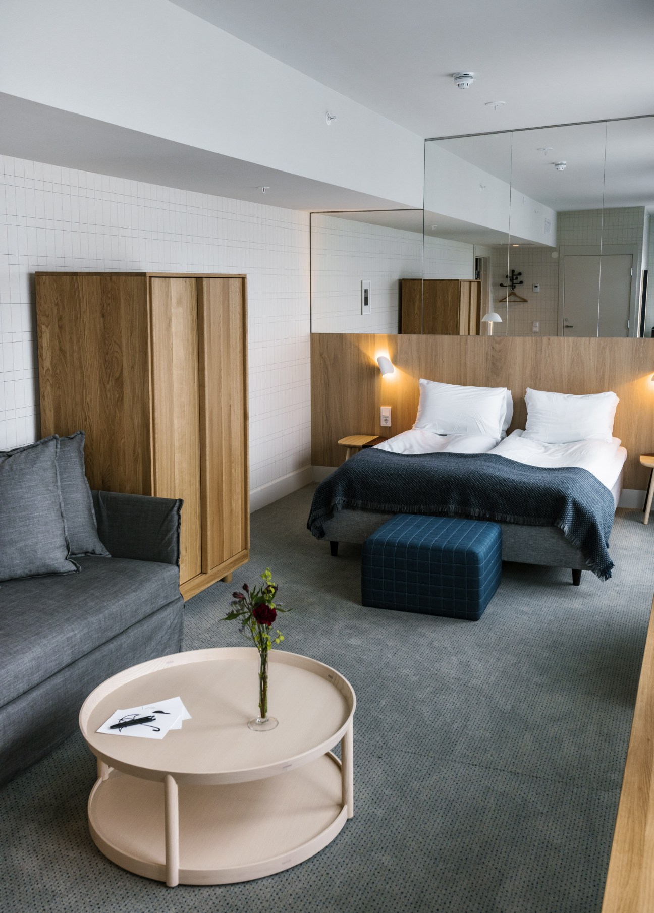 Modern hotel junior suite with wooden headboard, white pillows, dark blanket, and round cream table with flowers.
