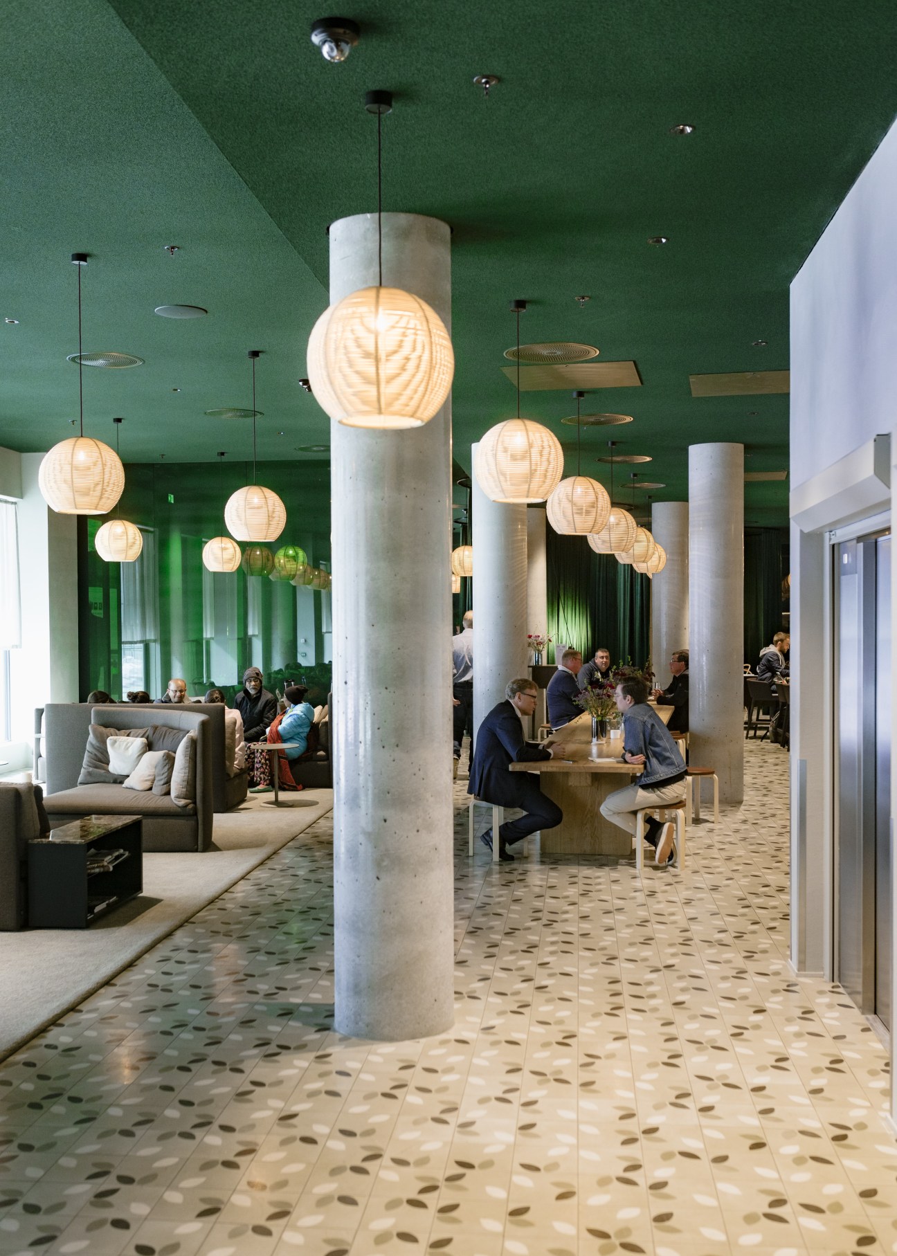Spacious lobby at Zander K hotel with green ceiling, pendant lights, concrete columns, and guests dining at wooden tables.