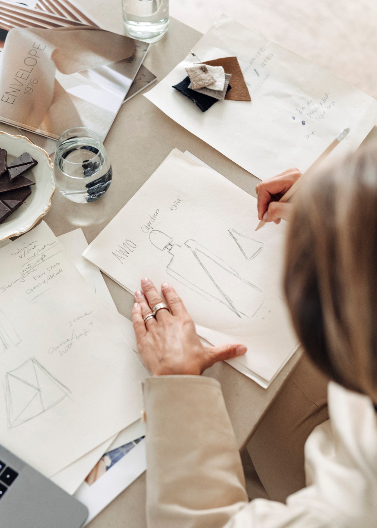 Designer sketching fashion design on white paper at a work table with fabric swatches and water glasses.
