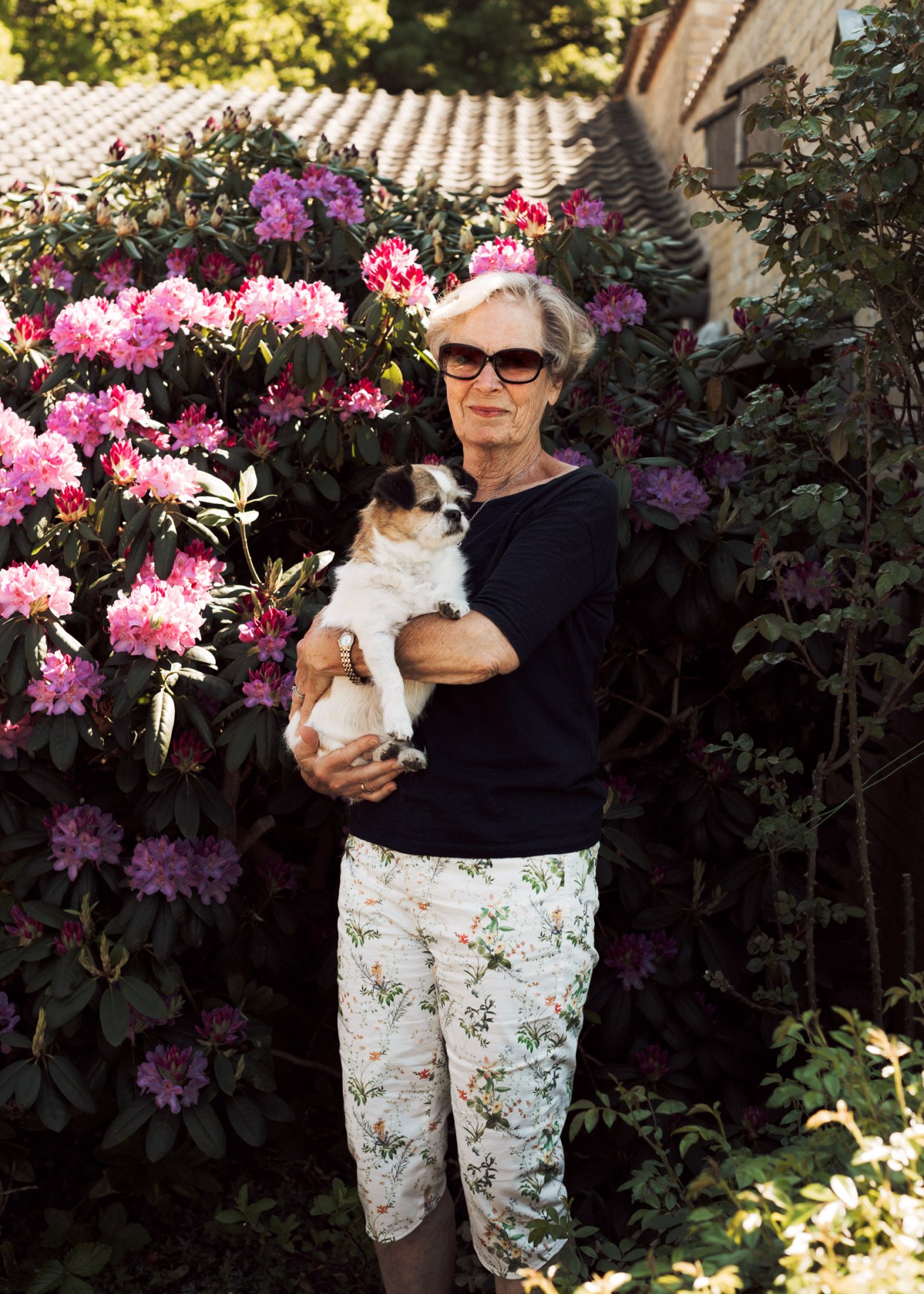 Resident Lissie Christoffersen holding her dog Oskar in a garden surrounded by pink rhododendron flowers.