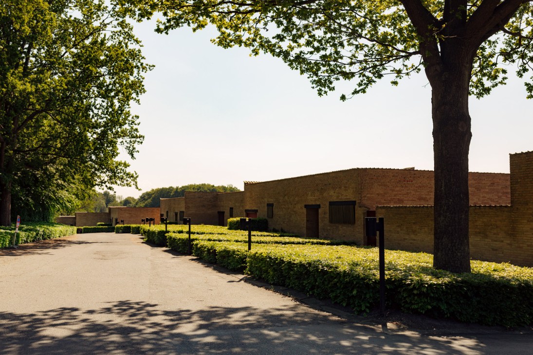 Brick buildings of the Fredensborg Houses nestled among oak and beech trees along a paved pathway.