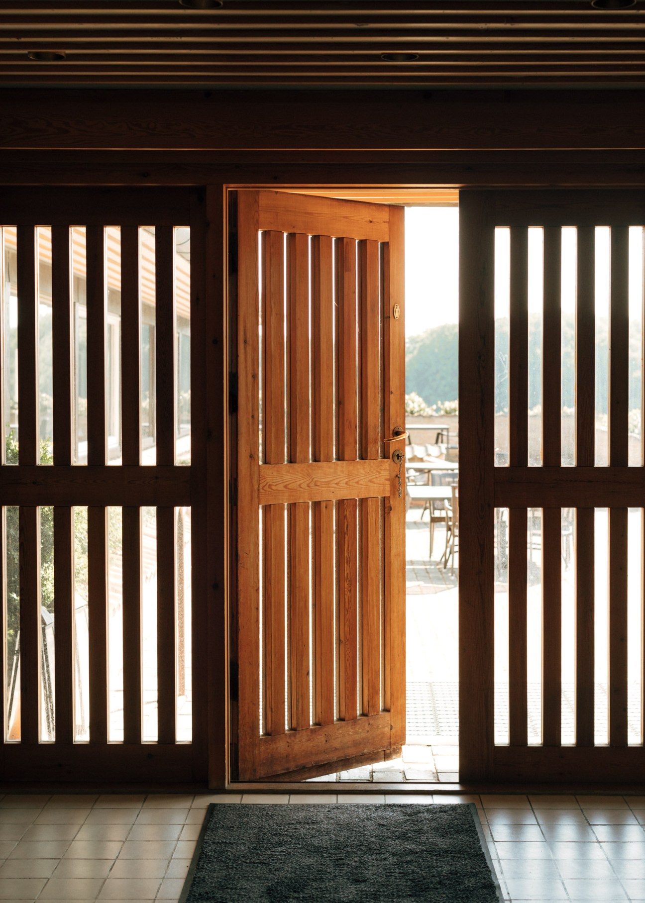 Wooden door with vertical slats opens onto a patio with mountain views beyond, flanked by dark wooden slotted screens.