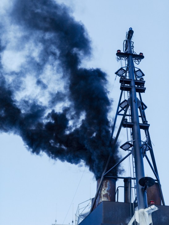 A tall ship mast with communication equipment silhouetted against a sky with dark smoke billowing from the vessel's funnel.